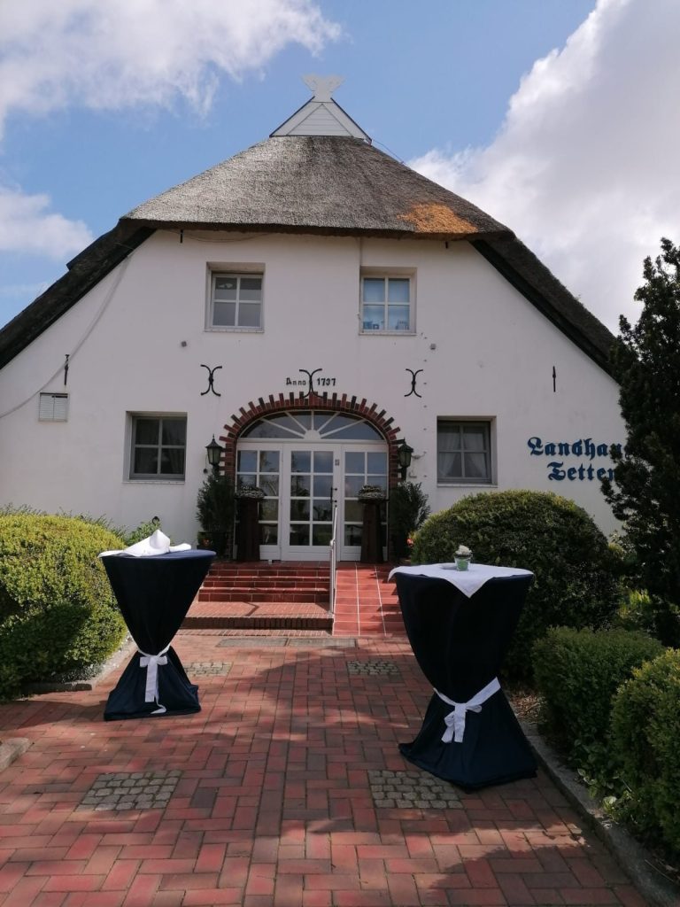Landhaus Tettens 2025 Helles Gebäude mit spitzem Dach, Eingangsbereich und dekorierten Stehtischen.