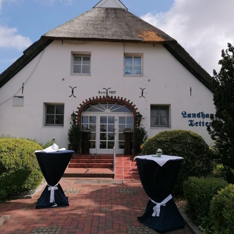 Landhaus Tettens 2025 Helles Gebäude mit Reetdach und zwei gedeckten Stehtischen im Vordergrund.